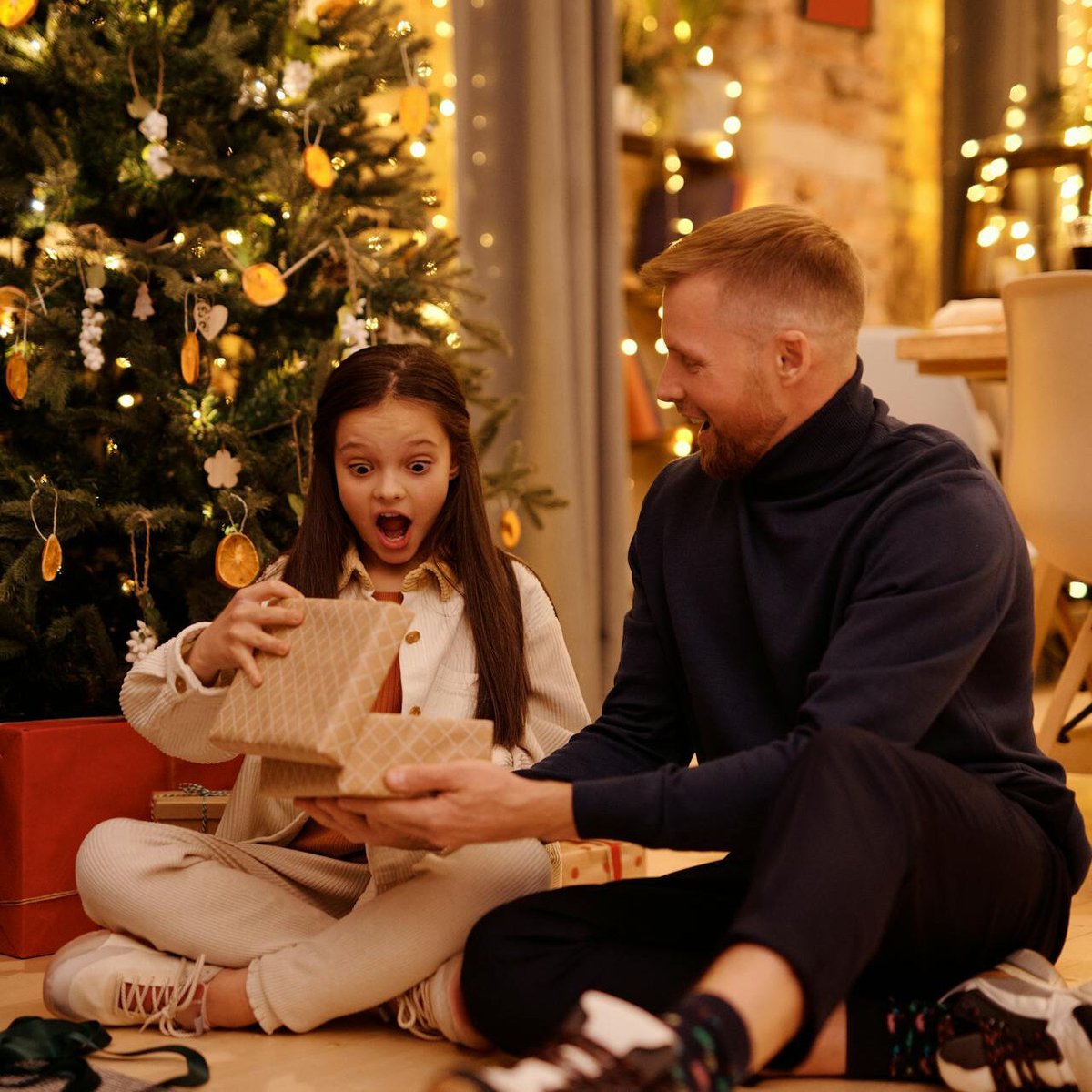 Девочка с удивлением смотрит в коробку с подарком, сидя рядом с папой у ёлки
